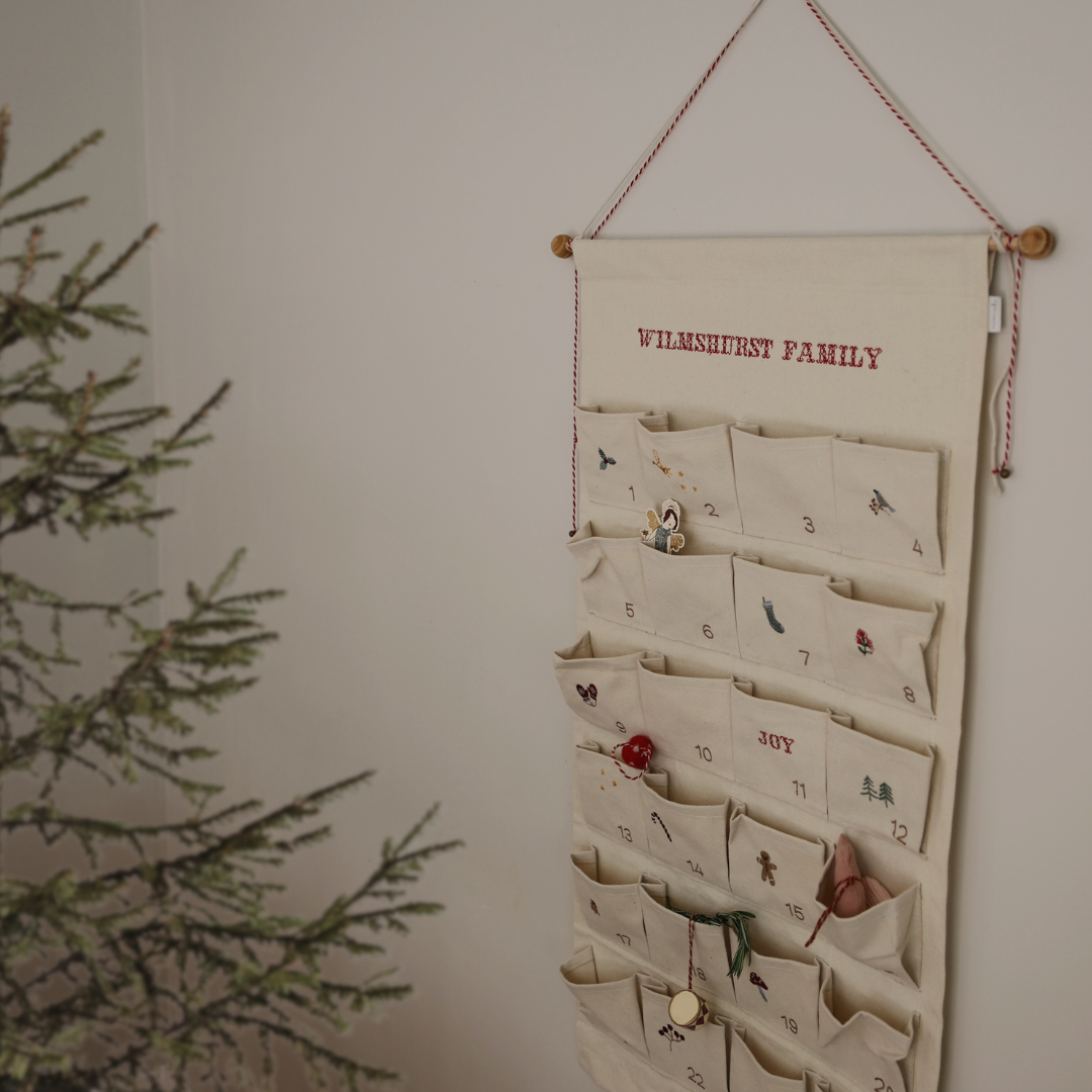 Embroidered advent calendar with personalized family name, made of ivory cotton canvas with numbered pockets and decorative red and white hanging string with tiny bells.