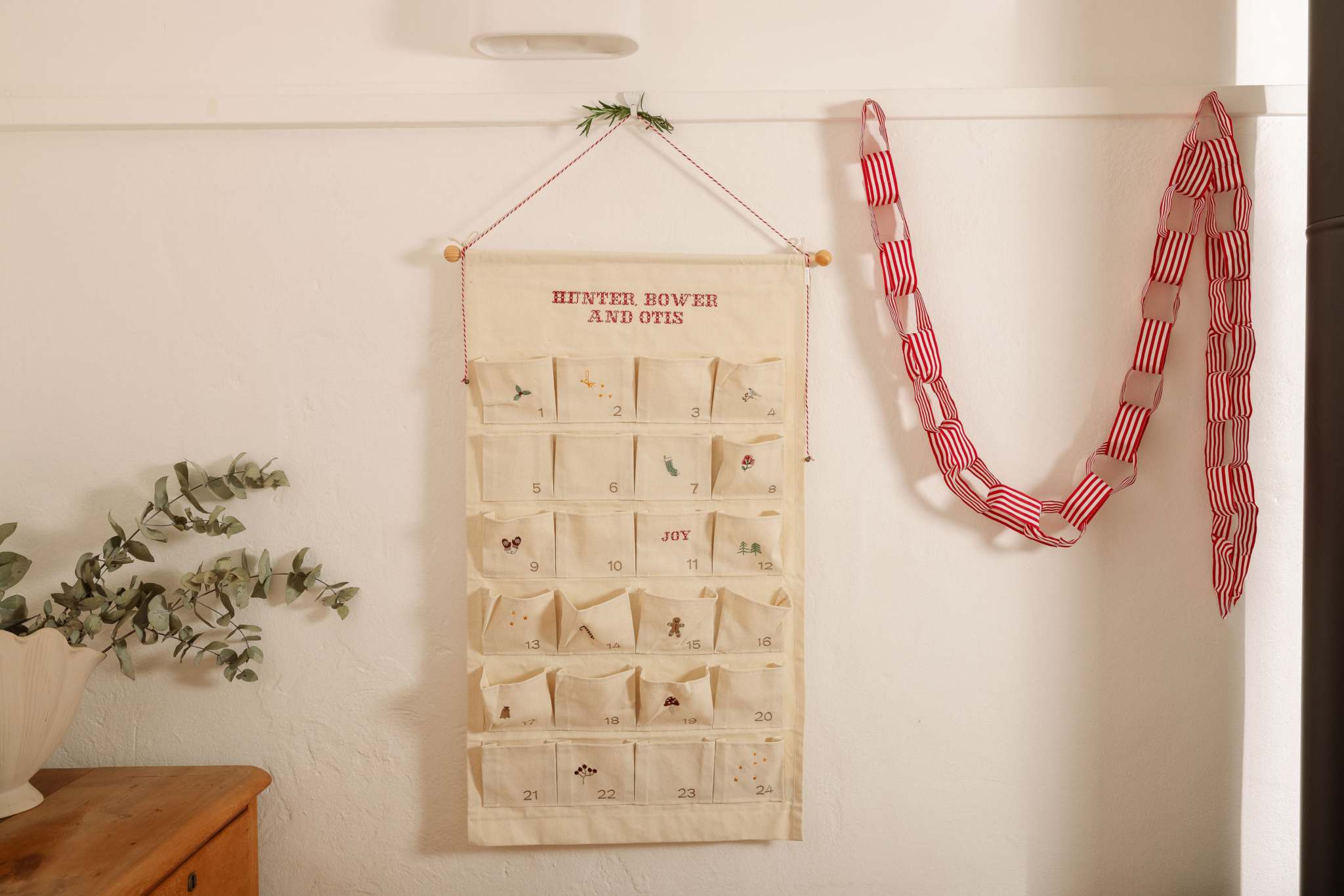 Embroidered ivory cotton canvas advent calendar with personalized Nordic Cross Stitch design and decorative red and white hanging string with tiny bells