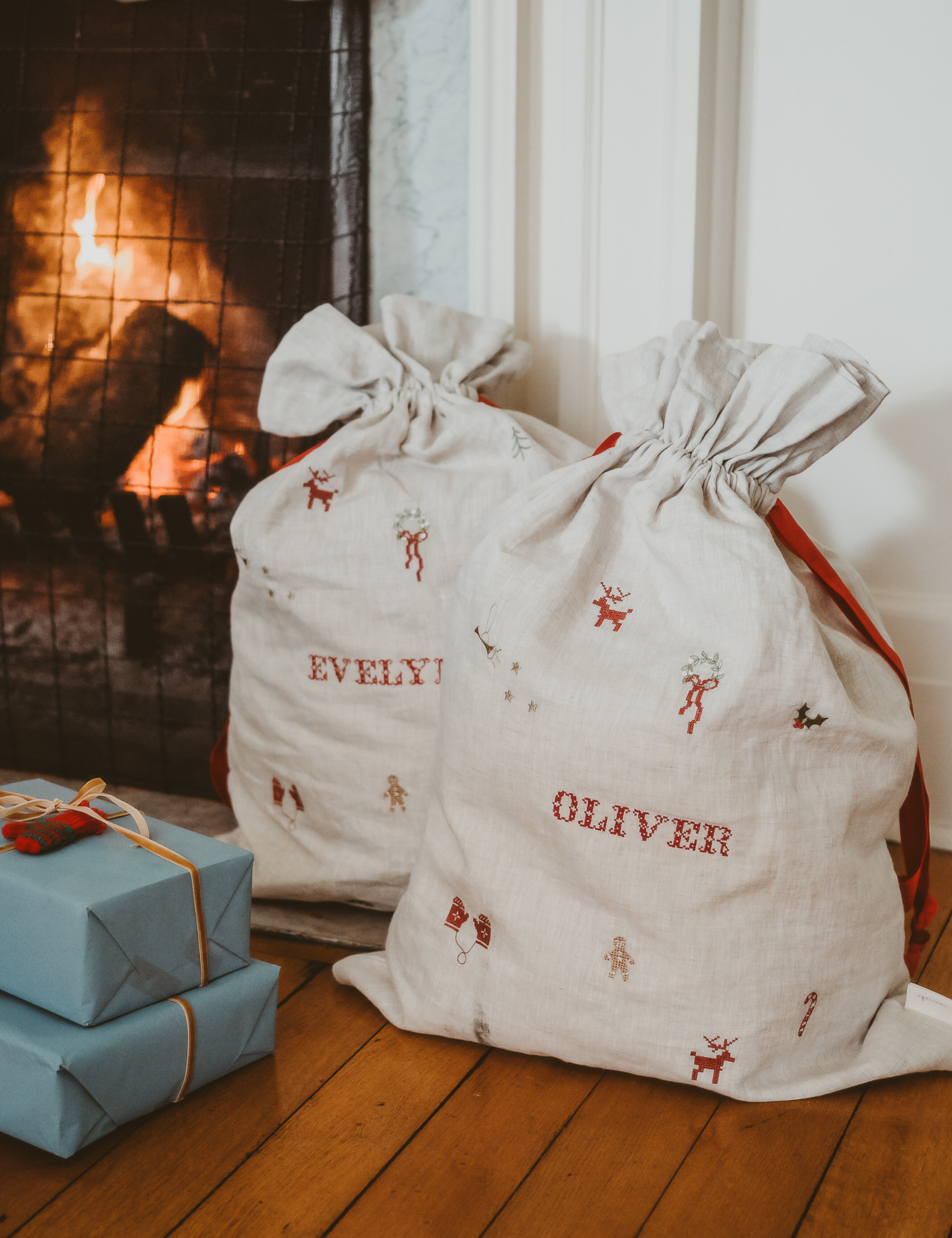 Natural linen Santa sacks with embroidered holiday motifs and personalized names Evelyn and Oliver by fireplace