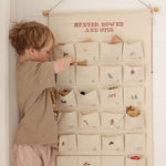 Child interacting with ivory cotton canvas embroidered advent calendar featuring personalized red text and 24 numbered pockets with whimsical designs and tiny bells on hanging string