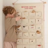Child interacting with ivory cotton canvas embroidered advent calendar featuring personalized red text and 24 numbered pockets with whimsical designs and tiny bells on hanging string