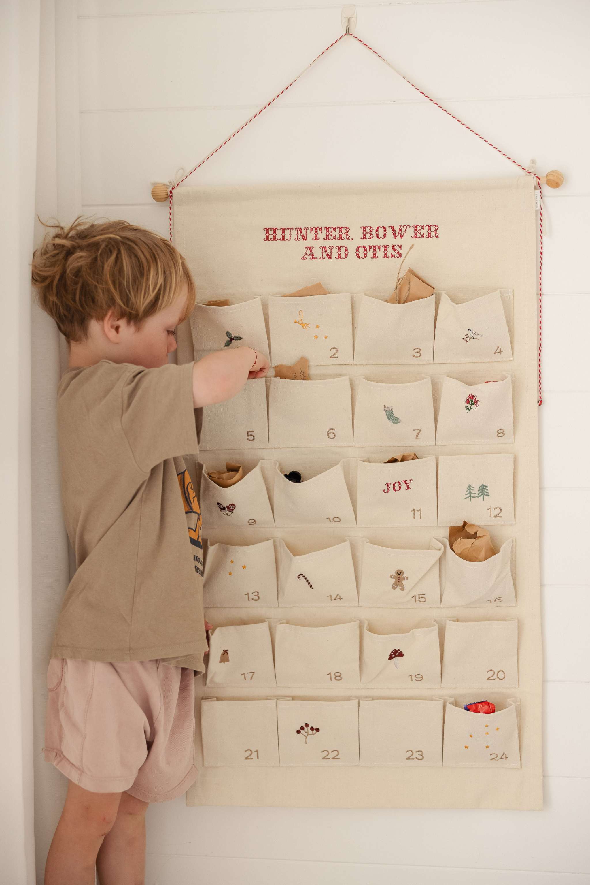 Child interacting with ivory cotton canvas embroidered advent calendar featuring personalized red text and 24 numbered pockets with whimsical designs and tiny bells on hanging string