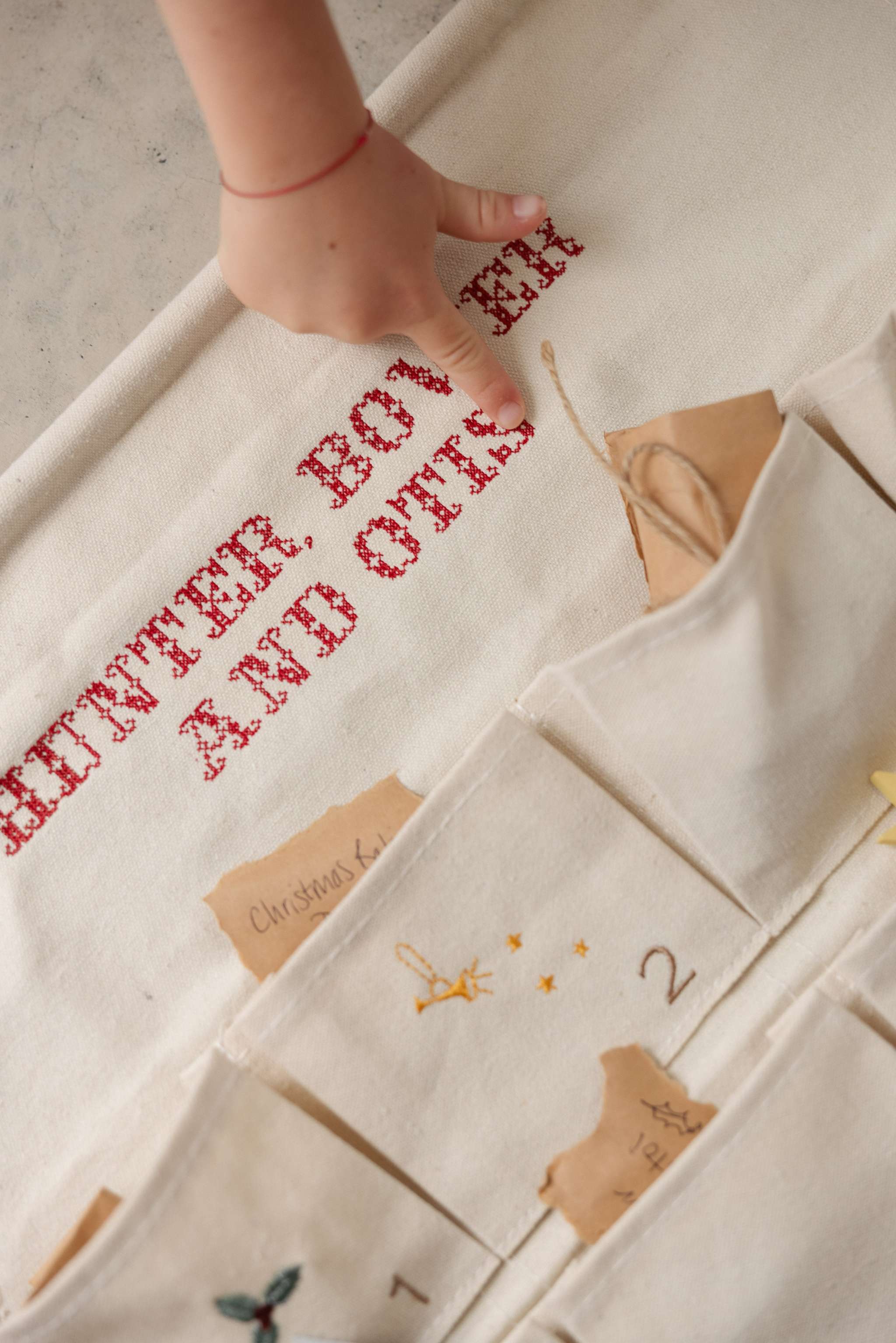 Handcrafted ivory cotton canvas embroidered advent calendar with whimsical designs and personalized vintage-style red cross stitch text and pockets.